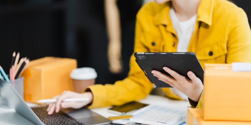Une femme avec une veste jaune tape sur son ordinateur et porte une tablette tactile